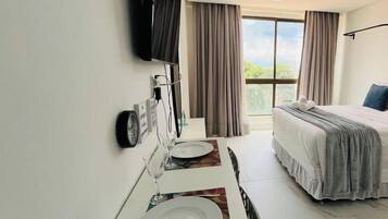 Una habitación de hotel con cama, televisión montada en la pared, una mesa de comedor con plato y vaso, y una ventana con cortinas.