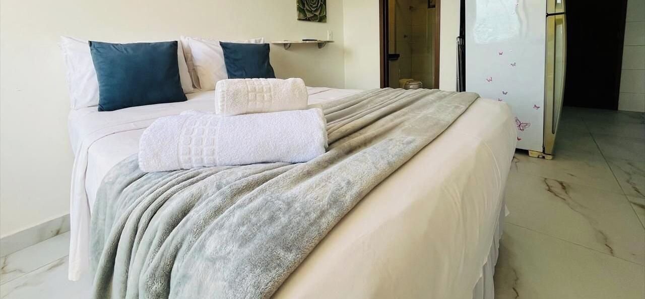 A hotel room with a bed, pillows, a blanket, a refrigerator, and a bathroom door.