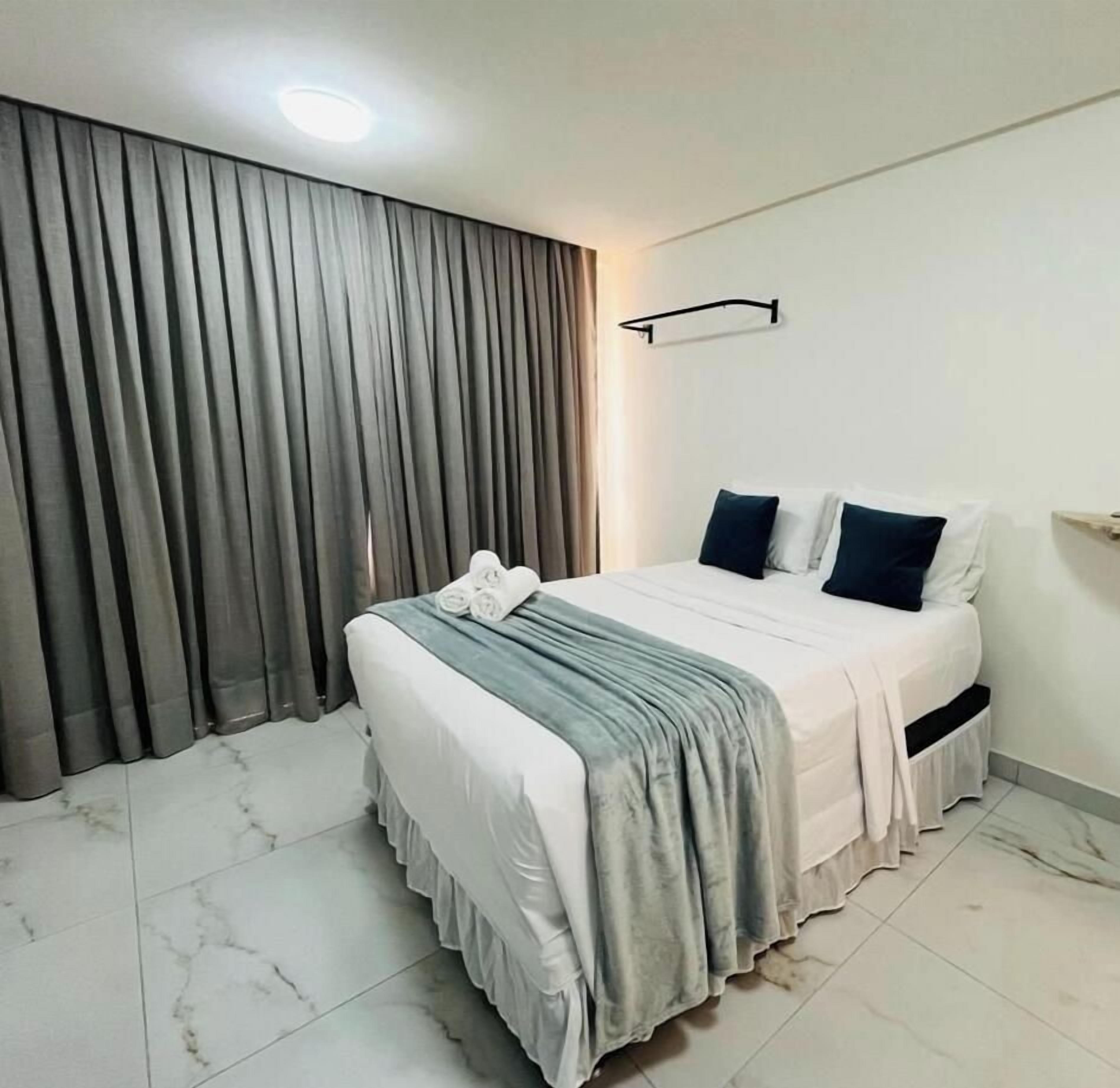 Una habitación de hotel con cama, cortinas, cabecero e iluminación empotrada en la pared.