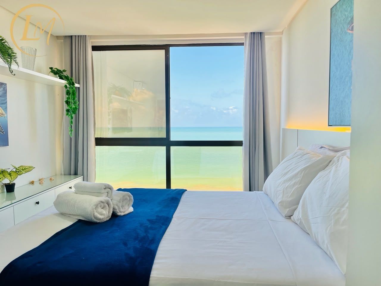 Een hotelkamer met een bed, een blauwe deken, witte kussens, een nachtkastje, een spiegel en uitzicht op zee.