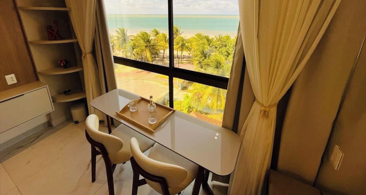 A room with a table, two chairs, a window with a view of the beach, and a shelf with items.