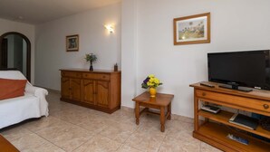 Ein Wohnzimmer mit einer Couch, einem Holzkabinett, einem kleinen Tisch mit Blumen, einem Fernseher und einem gerahmten Bild an der Wand.