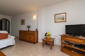 Ein Wohnzimmer mit einer Couch, einem Holzkabinett, einem kleinen Tisch mit Blumen, einem Fernseher und einem gerahmten Bild an der Wand.