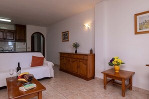 Ein Wohnzimmer mit einer Couch, einer Holzkommode, einem kleinen Tisch mit einer Vase und einer Küchenzeile im Hintergrund.