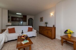A living room with a sofa, a wooden coffee table with a book and a bottle, and a dining area with a wooden cabinet. - Paloma Beach 16 - One Bed (Los Cristianos)