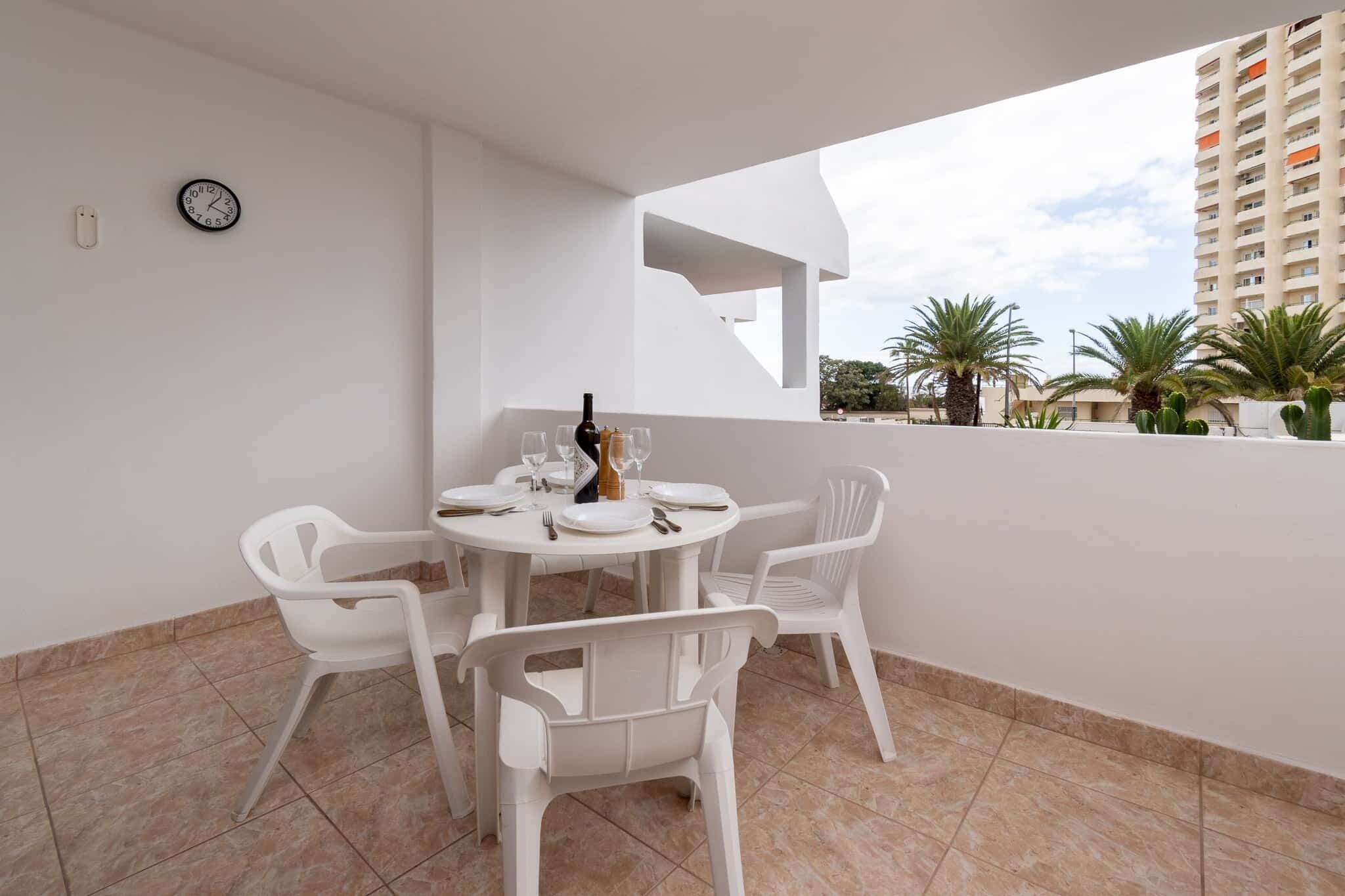 A white table with four chairs, a bottle, and glasses on a balcony with a view of palm trees and a building.