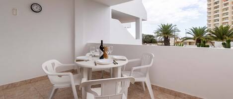A white table with four chairs, a bottle, and glasses on a balcony with a view of palm trees and a building.