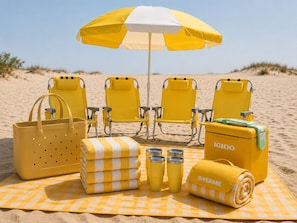 Een strandopstelling met vier gele stoelen, een gele parasol, een gele koelbox, een gele strandtas, een stapel gele en witte handdoeken en drie gele bekerbekers op een geruit strandkleed.