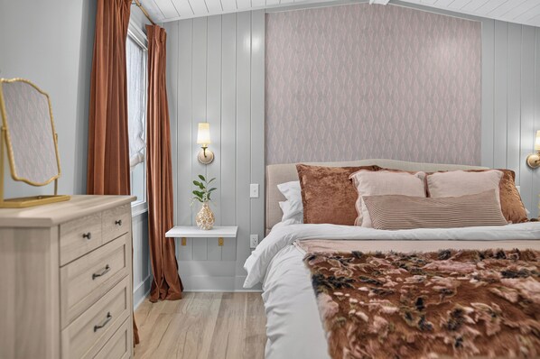 A bedroom with a bed, a dresser, a mirror, a wall-mounted lamp, and a patterned wall.