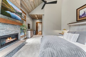Ein Schlafzimmer mit Kamin, einem Bett mit grauer Bettwäsche, einem Deckenventilator und einem Gemälde an der Wand.