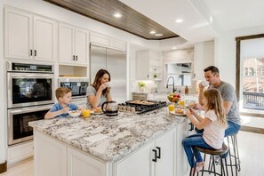 A modern kitchen with white cabinetry, a marble island, and a central island with breakfast items. - 🏔️ Leika Haus | Riverfront Luxury ♨️ Hot Tub + Barrel Sauna 🎬 Theater Lounge | Sleeps 19 (Keystone)