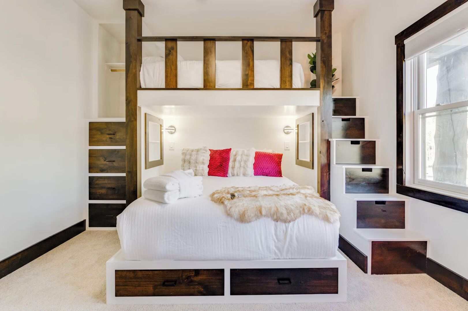 A bunk bed with a white mattress, red and white pillows, and a fur throw. There are wooden storage drawers under the bed and a small plant on the windowsill.