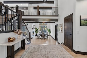 A spacious interior with a high ceiling, a staircase, a dining area, and a view through a large window. - 🏔️ Leika Haus | Riverfront Luxury ♨️ Hot Tub + Barrel Sauna 🎬 Theater Lounge | Sleeps 19 (Keystone)