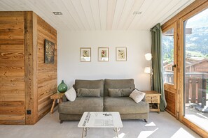 A cozy room with a wooden accent wall, a grey sofa with cushions, a small table with a sign, and framed artwork on the wall. - Charming Duplex – Les Confins: Mountain View & Balcony (La Clusaz)