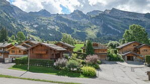A cluster of wooden mountain cabins with stone foundations, surrounded by lush greenery and a backdrop of rugged mountains.