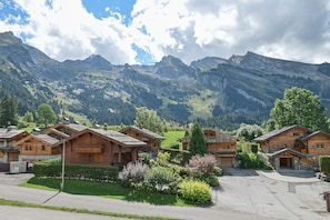A cluster of wooden mountain cabins with stone foundations, surrounded by lush greenery and a backdrop of rugged mountains.