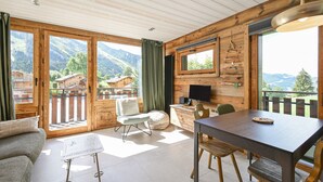 A wooden interior with a dining area, a TV mounted on the wall, and a view of mountains through large windows.