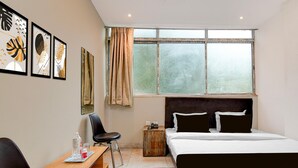 A hotel room with a bed, a desk with a chair, a window with curtains, and artwork on the wall. - Deluxe The Royal Orbit (Barasat)