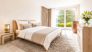 A bedroom with a large bed, a nightstand with a lamp, a dresser, and a view of greenery outside. - Garden Oasis Apartment (Prague 2)