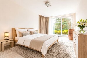A bedroom with a large bed, a nightstand with a lamp, a dresser, and a view of greenery outside. - Garden Oasis Apartment (Prague 2)