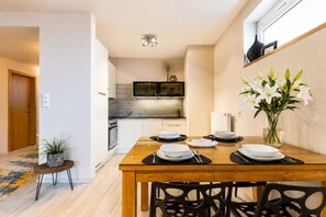 A dining area with a wooden table set for four, a vase of white flowers, and a view into the kitchen with a modern stove and microwave. - Garden Oasis Apartment (Prague 2)