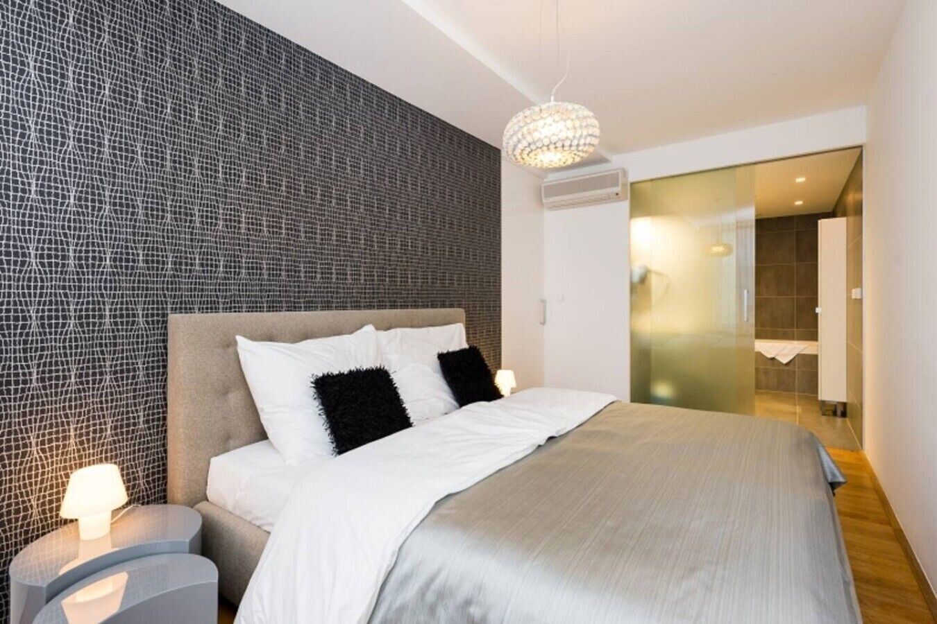 A modern bedroom with a large bed, a textured wall, a chandelier, and a bathroom visible through a glass door.