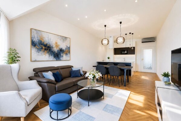 Una sala moderna con sofá, reposapiés y una mesita de centro, un comedor con sillas, y una cocina al fondo.