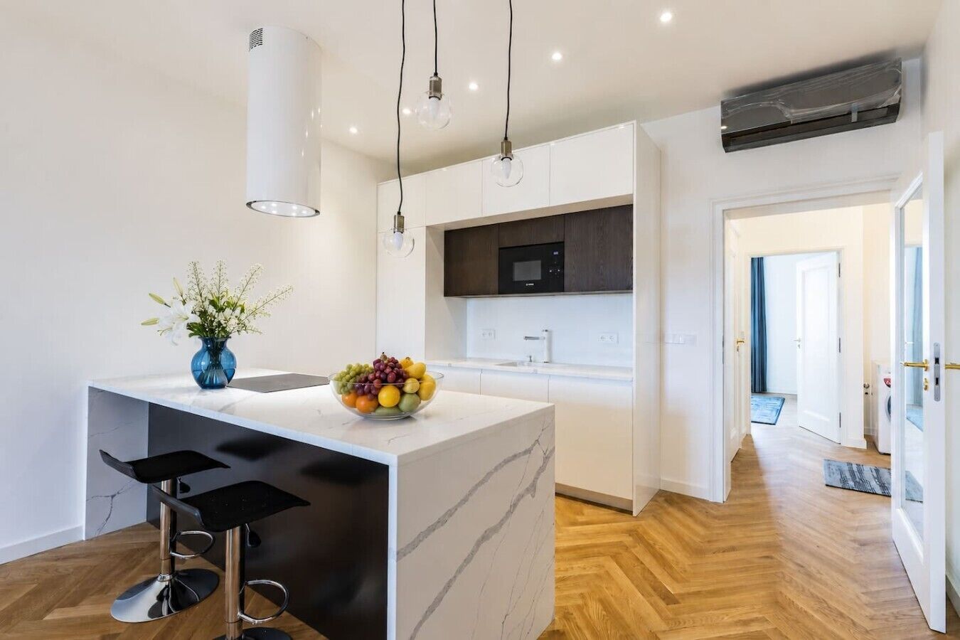 A modern kitchen with a marble island, black bar stools, a fruit bowl, and pendant lighting.