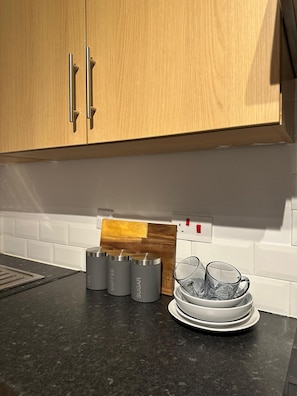 A kitchen with wooden cabinets, a black countertop, stainless steel containers labeled tea, coffee, and sugar, a wooden cutting board, and stacked white plates. - Modern 1-Bedroom | Free Parking | Hospital Bolton (bolton)