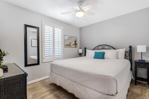 A bedroom with a bed, a ceiling fan, a window with blinds, a painting, and a nightstand with a lamp. - 🏖️ Grand Caribbean 423 | 2BR Orange Beach | Steps to Beach + Pool + Fitness (Orange Beach)