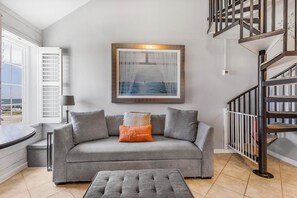A living room with a grey sofa, a small ottoman, a framed picture of a seascape, a staircase, and a window with blinds. - 🏖️ Grand Caribbean 423 | 2BR Orange Beach | Steps to Beach + Pool + Fitness (Orange Beach)