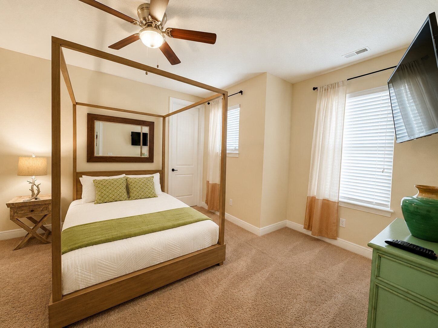 Un lit à baldaquin avec un ventilateur de plafond, une télévision et un vase vert posé sur une commode.