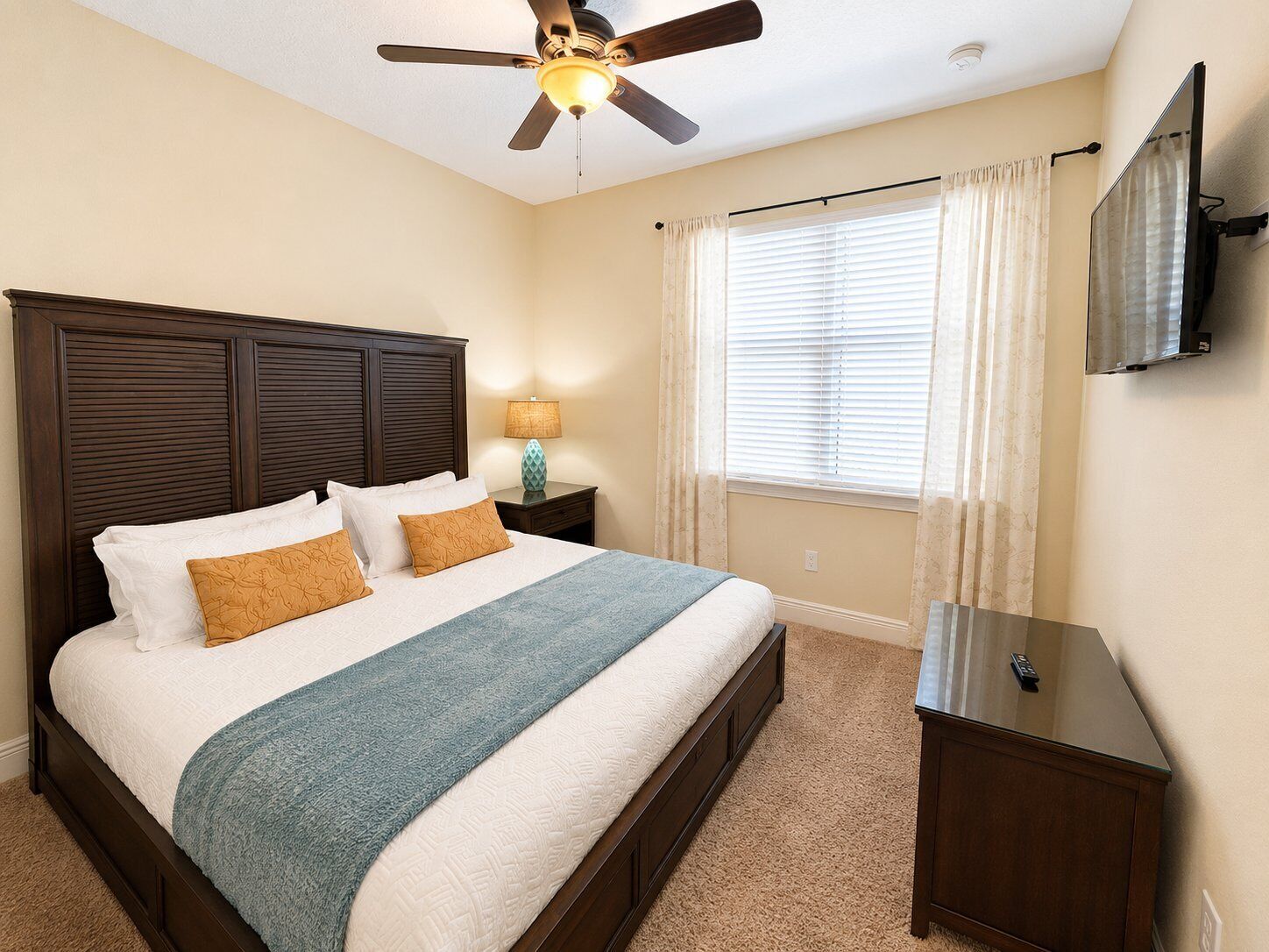 A bedroom with a bed, a nightstand, a lamp, a ceiling fan, a window with curtains, and a TV mounted on the wall.