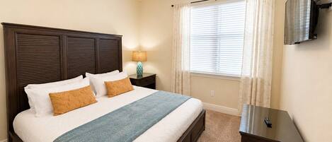 A bedroom with a bed, a nightstand, a lamp, a ceiling fan, a window with curtains, and a TV mounted on the wall.