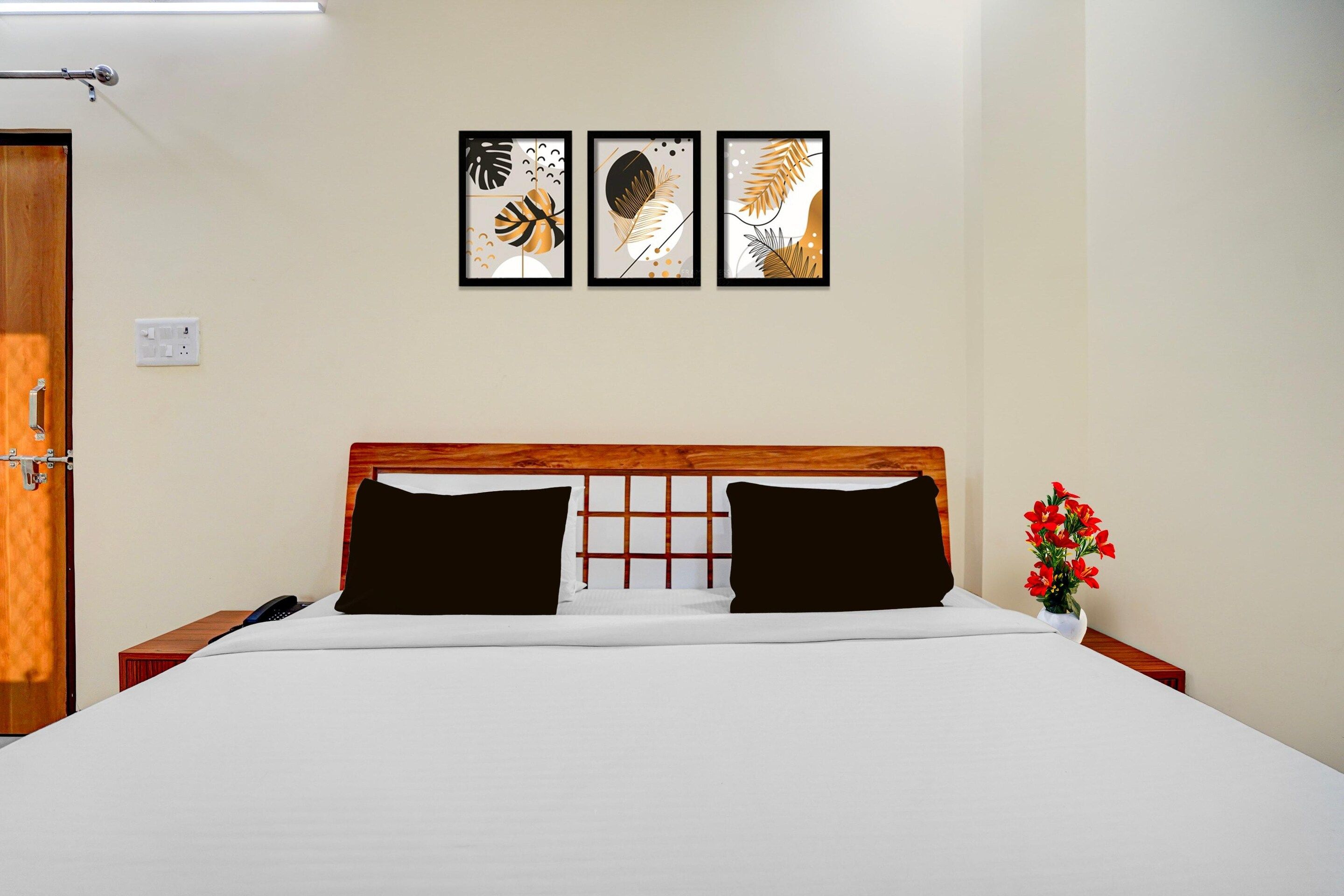 A hotel room with a wooden headboard, a bed with white bedding, three framed artworks on the wall, and a vase with red flowers on the nightstand.