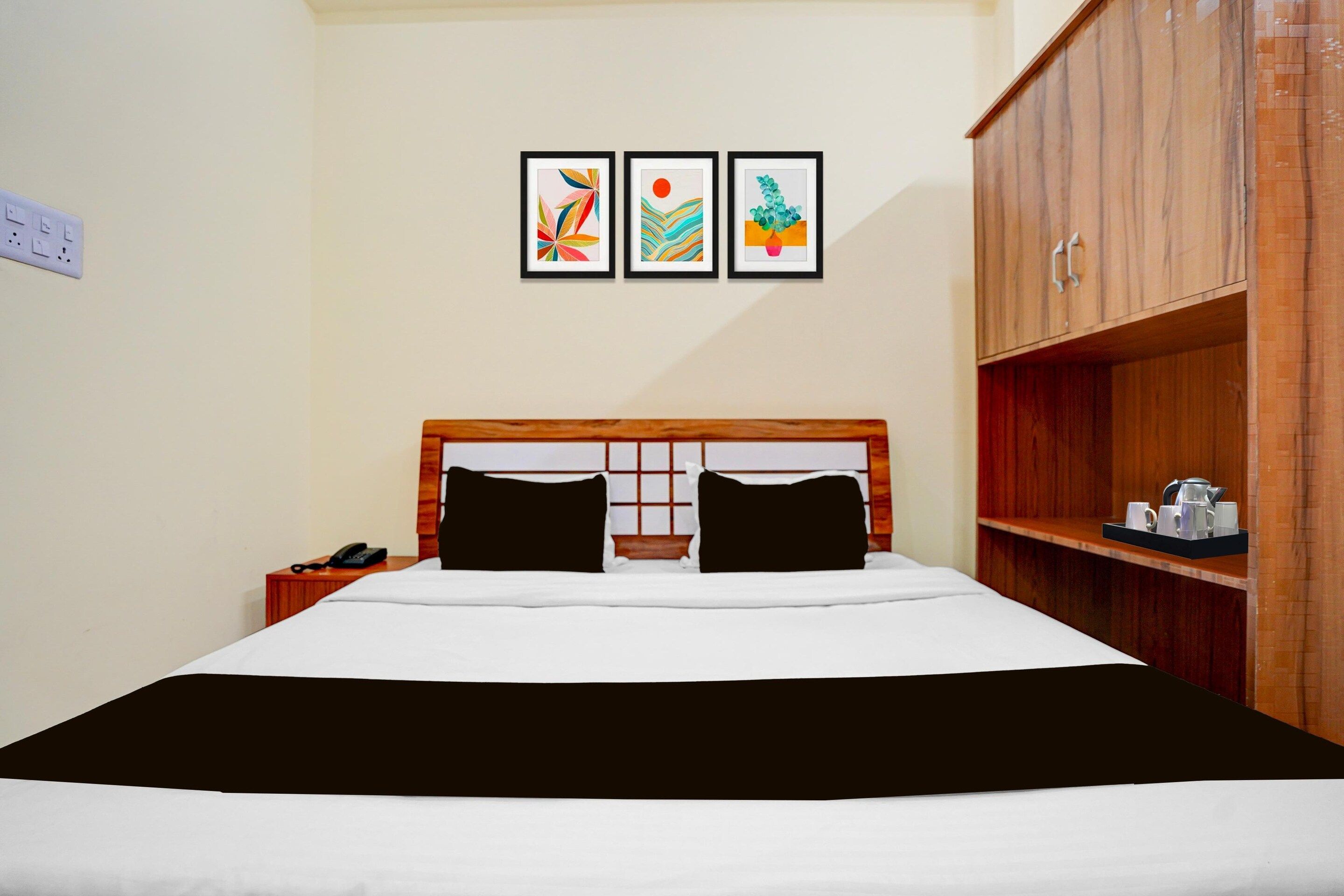 A hotel room with a bed, a bedside table with a phone, and a wardrobe. There are three framed artworks on the wall above the bed.