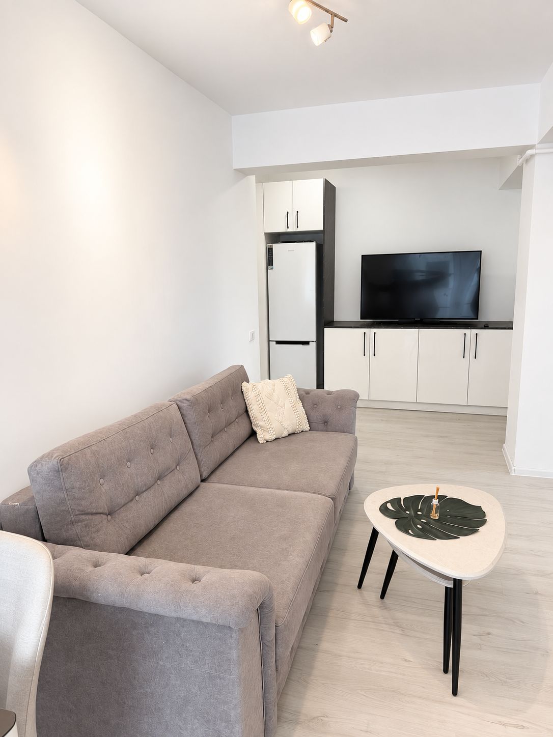 A modern living room with a grey sofa, a coffee table, a flat-screen TV, and a kitchen area with cabinets and a refrigerator.