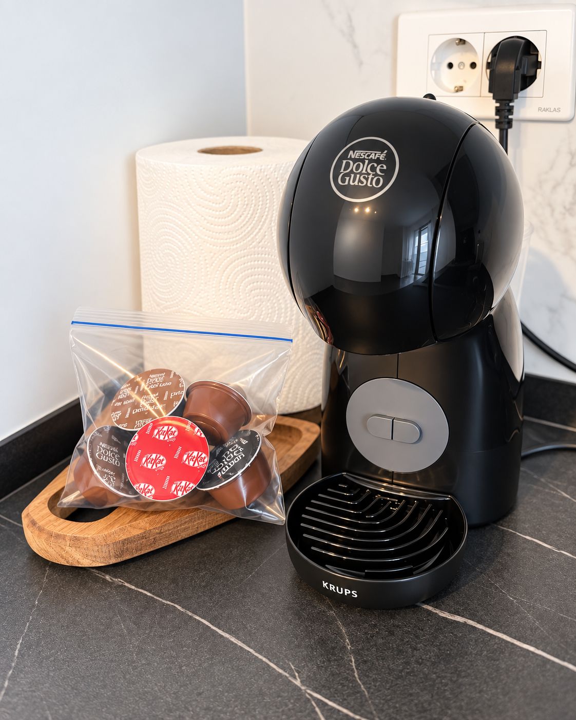 Eine Nescafé Dolce Gusto Kaffeemaschine mit einem Krups-Kaffeekännchen auf einer Küchenarbeitsplatte, eine Rolle Küchenpapier und eine Packung Nescafé Dolce Gusto Kapseln.