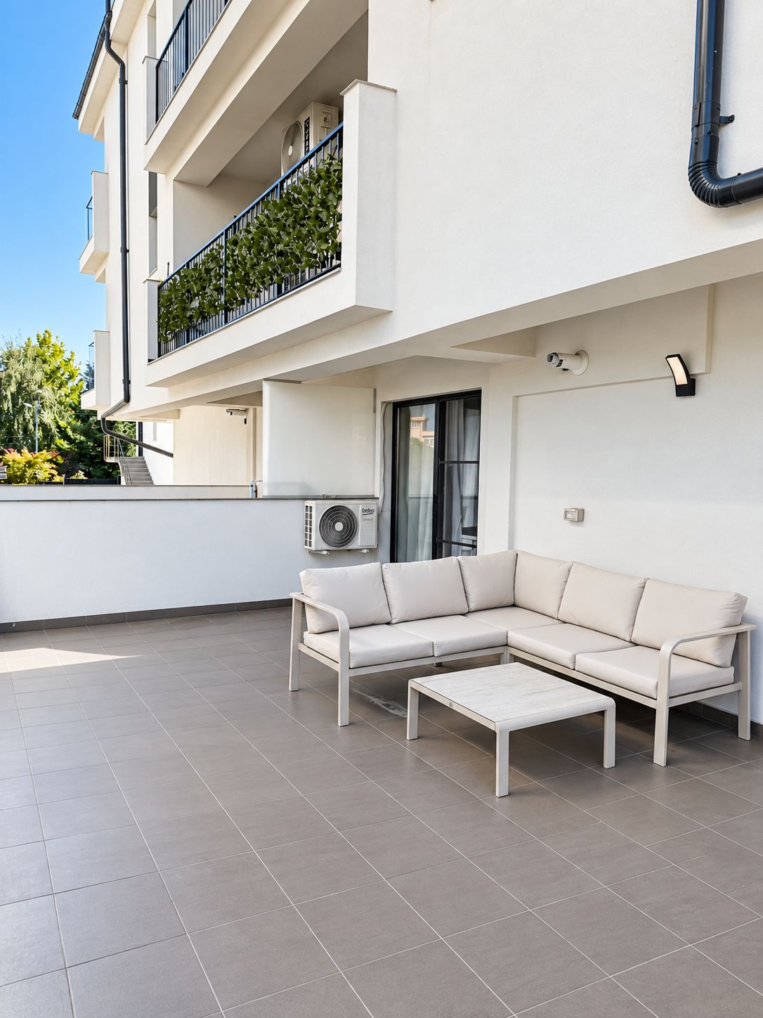 Ein moderner Außenbereich mit einer weißen Couch und passendem Tisch, einem Balkon mit schwarzer Geländer und einer Klimaanlage.