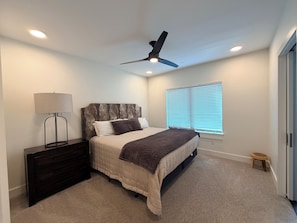 Ein Schlafzimmer mit Bett, Nachttisch, Lampe, Deckenventilator, Fenster mit Jalousien und einem kleinen Hocker.