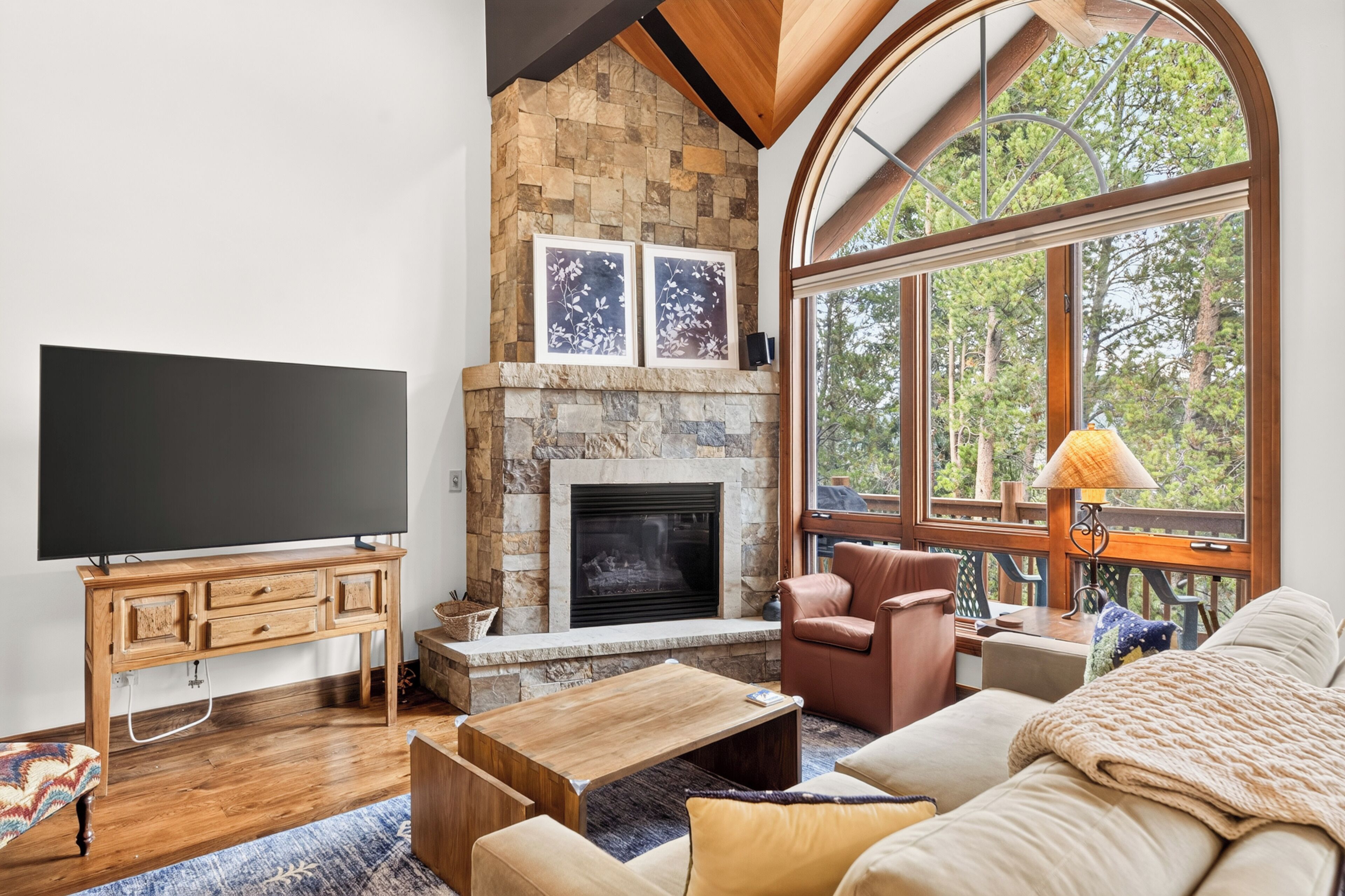 A living room with a stone fireplace, a television, and a large window with a view of trees.