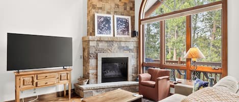 A living room with a stone fireplace, a television, and a large window with a view of trees.