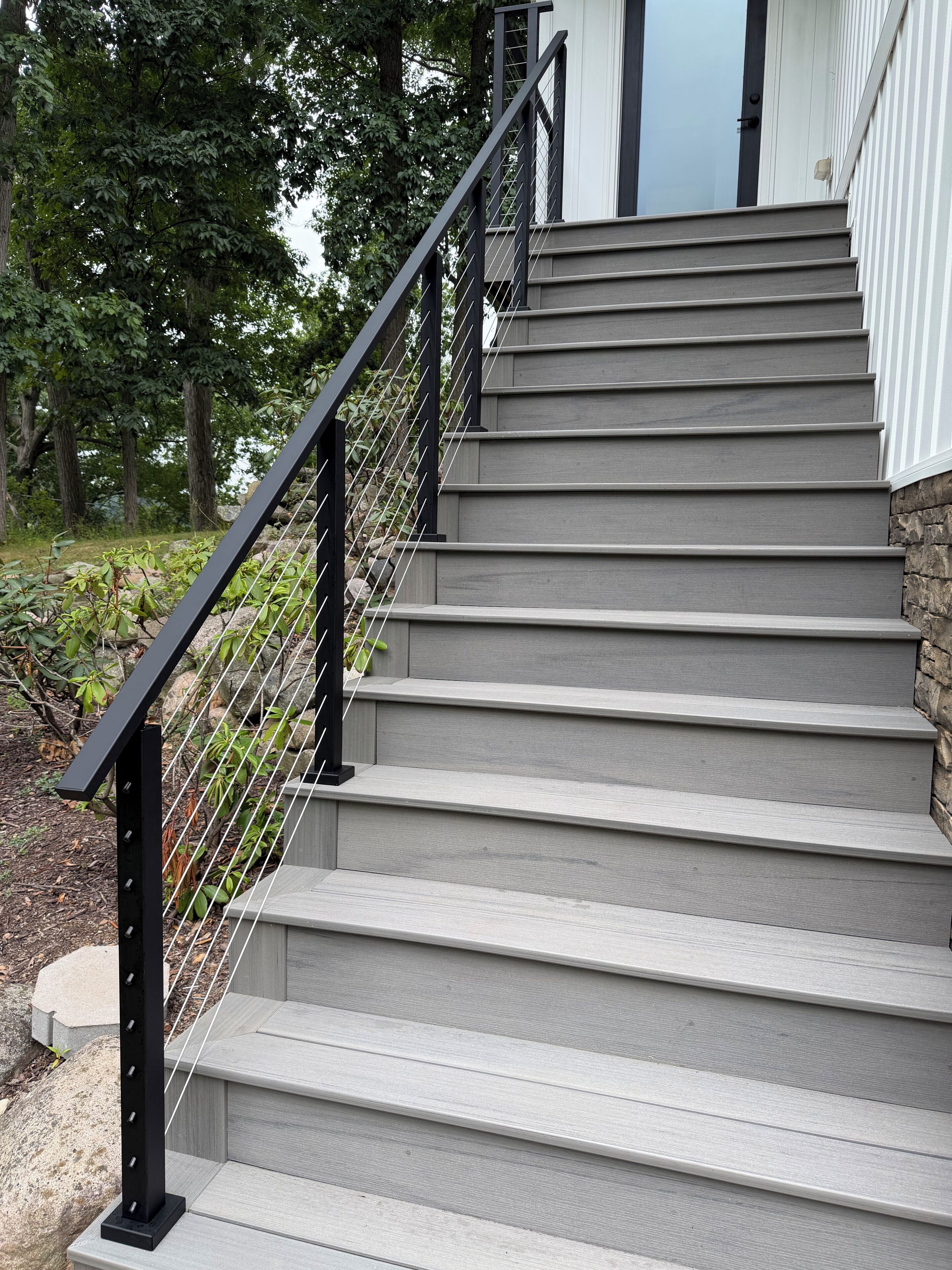 A set of grey wooden stairs with a black metal handrail and a stone wall on one side.
