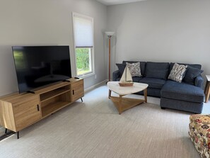 Ein Wohnzimmer mit einem Flachbildfernseher auf einer Holzeinrichtung, einem dunkelblauen Sofa mit gemusterten Kissen, einem kleinen runden Couchtisch und einem Fenster mit Jalousien.
