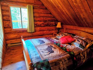 Ein Schlafzimmer in einer Blockhütte mit einem Bett, einem Fenster mit Vorhängen und einer Lampe.