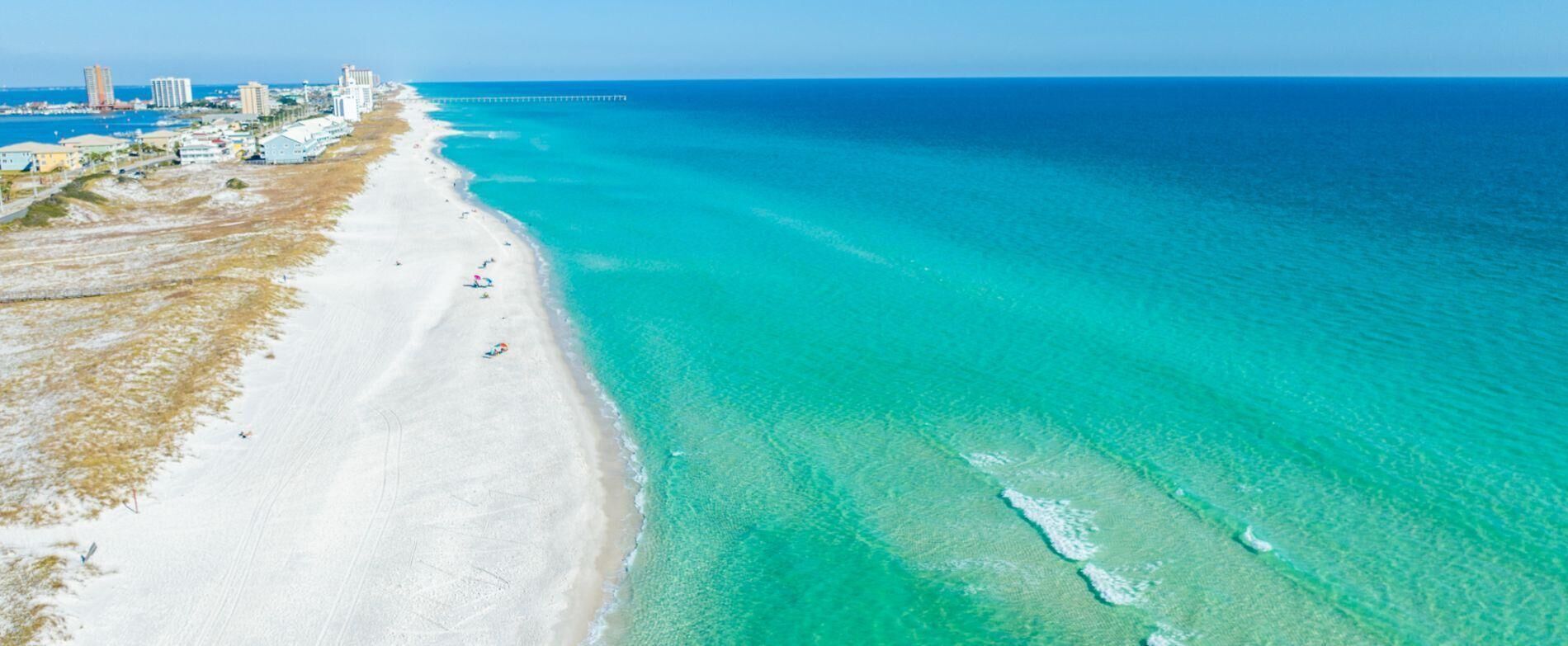 Una playa costera con vista despejada al océano, un muelle a lo lejos y edificios a lo largo de la costa.