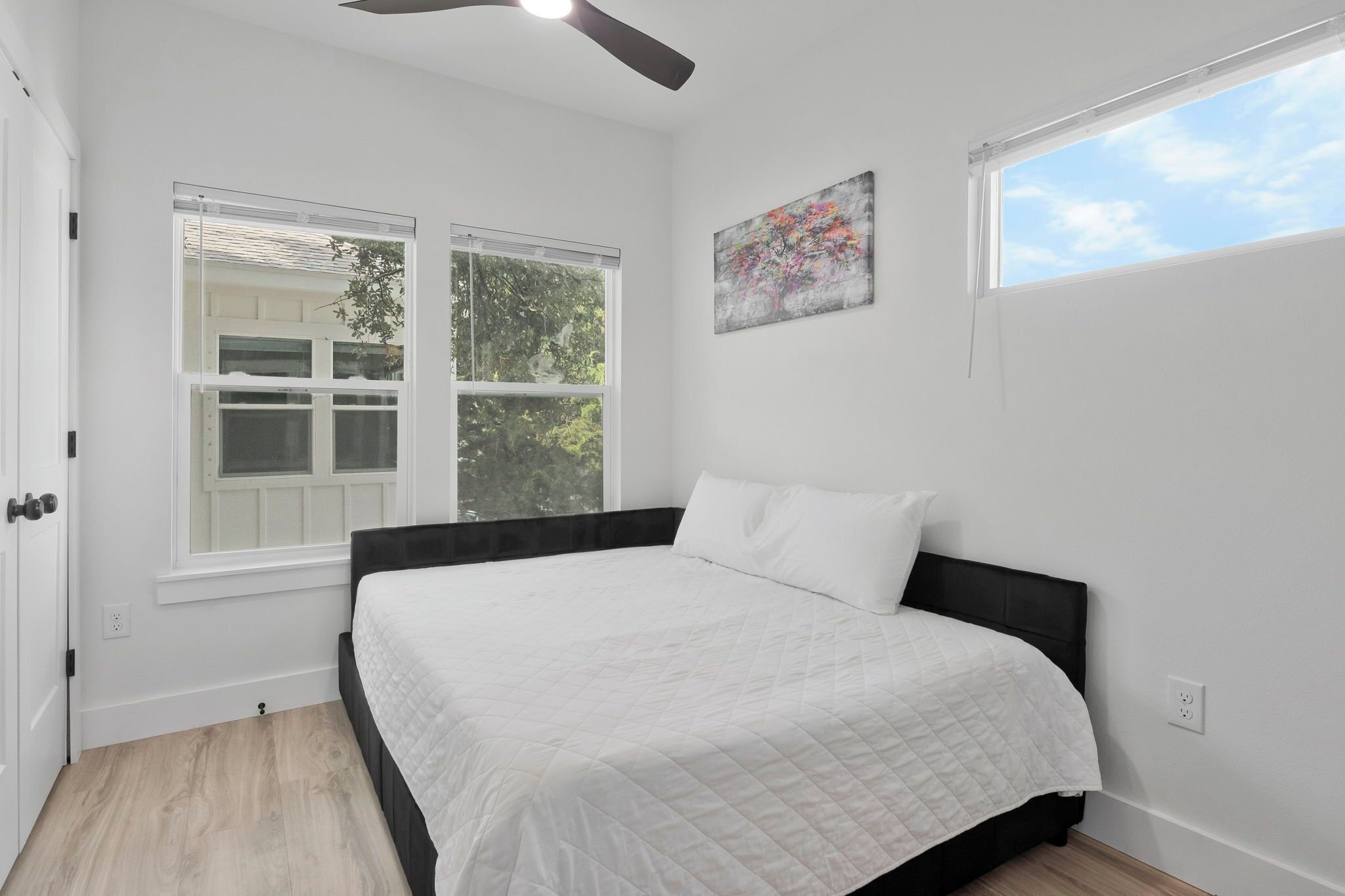 Une chambre à coucher avec un lit, deux fenêtres, un ventilateur de plafond et un tableau accroché au mur.