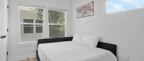 Une chambre à coucher avec un lit, deux fenêtres, un ventilateur de plafond et un tableau accroché au mur.