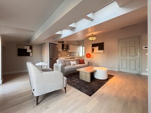 A modern living room with a sofa, armchair, coffee table, and a rug. There is a kitchen in the background with a window and a door. - Top-floor 1BR with private terrace (Istanbul)