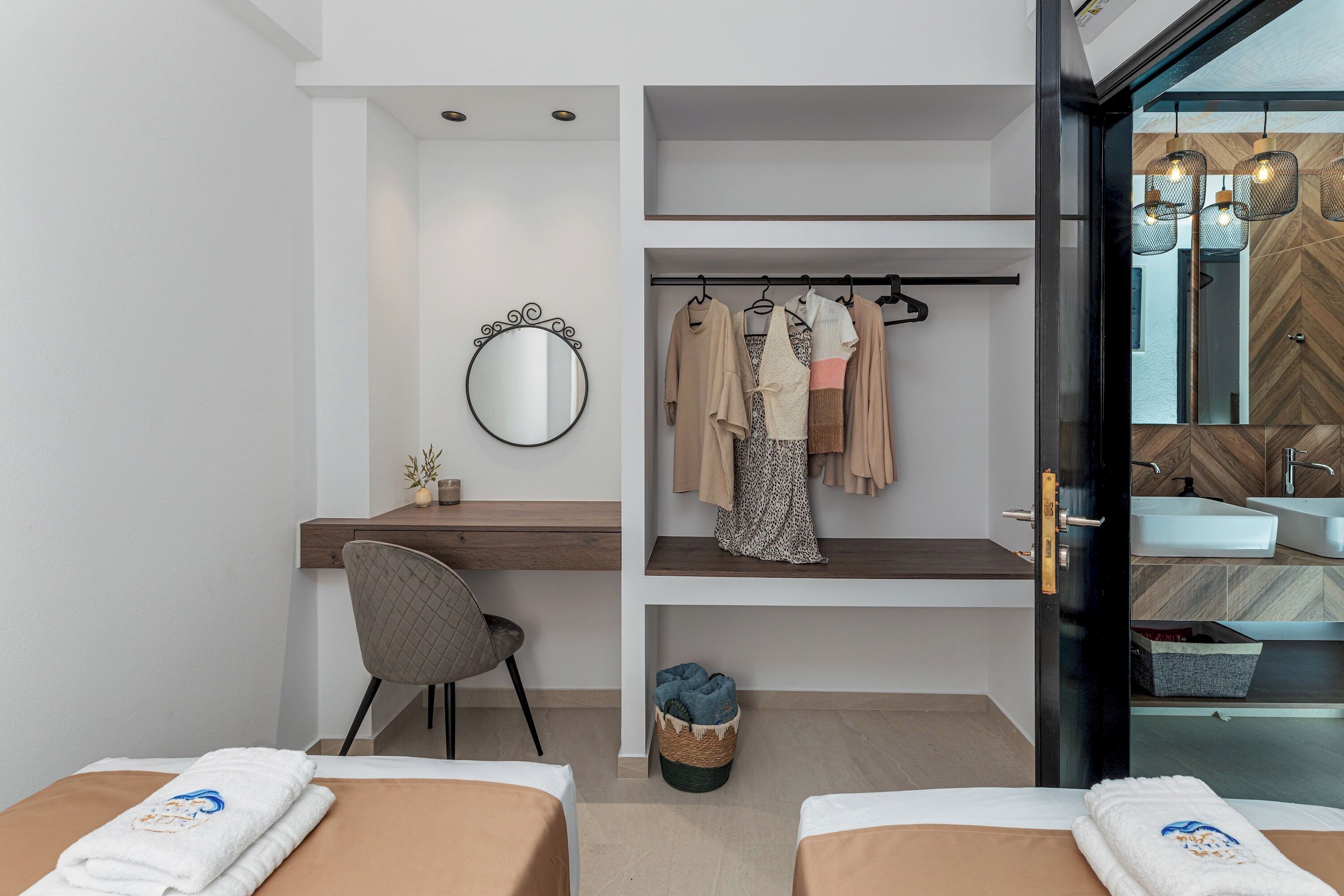 Une chambre d’hôtel moderne équipée d’un bureau, de deux lits, d’un miroir, d’un porte-vêtements et d’une salle de bain dont le dosseret est recouvert de carreaux disposés en motif chevrons.
